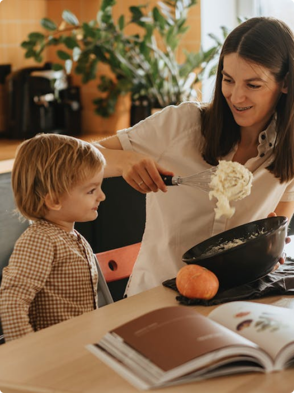 A mother and son are cooking together in a kitchen. The mother has dark hair and a white shirt. She is whisking the contents of a black bowl. The boy is watching.