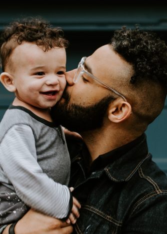 A man with dark hair and facial hair wearing glasses is giving the baby in his arms a kiss. The baby is smiling.