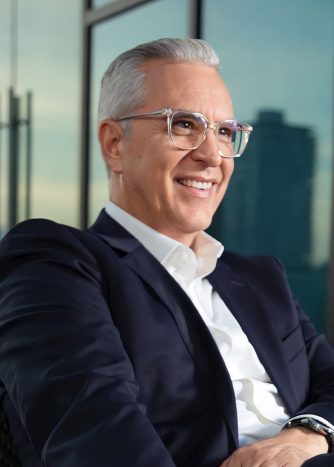 A man with silver hair and glasses wearing a suit is looking into the distance with a smile on his face.