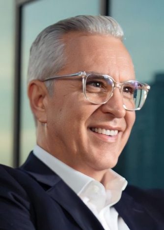 A man with silver hair and glasses wearing a suit is looking into the distance with a smile on his face.