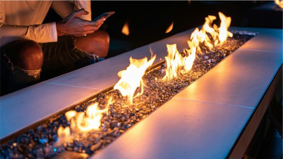 Close-up of a modern fire pit with flames rising from glass rocks, with a person seated nearby holding a phone.