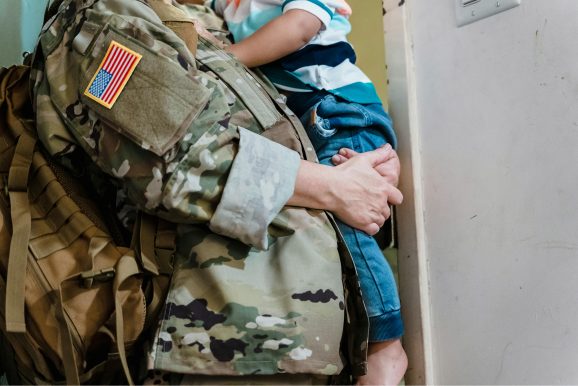 A U.S. military service member in camouflage uniform holds a small child in their arms, symbolizing family, service, and support for veterans.